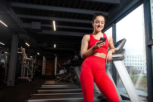 Une fille sportive en tenue de sport rouge utilise un smartphone dans la salle de sport et sourit