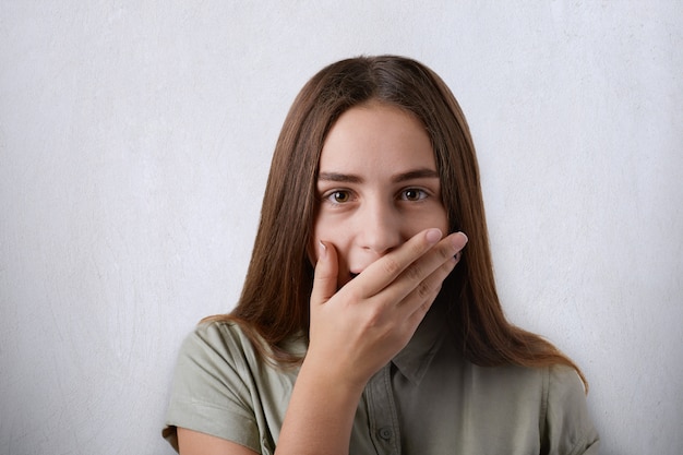 Fille surprise avec de longs cheveux raides et des yeux sombres couvrant sa bouche avec la main sur le gris