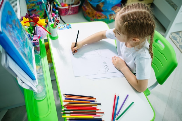 Une fille en T-shirt blanc est assise dans sa chambre à table et dessine avec des crayons de couleur