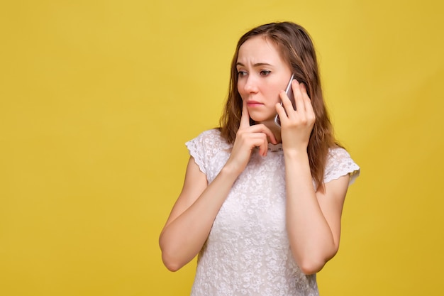 Une fille en T-shirt blanc et jean marron sur fond jaune parle avec inquiétude sur un smartphone.