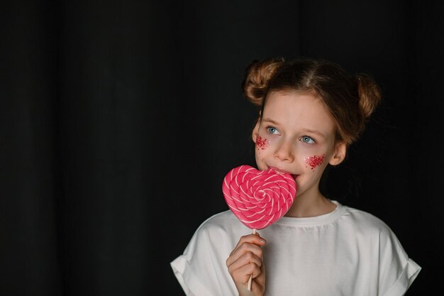 Une fille en T-shirt blanc mange une sucette en forme de cœur sur un fond noir.