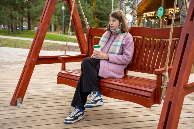 Fille avec une tasse de café en papier se balance sur un banc dans le parc