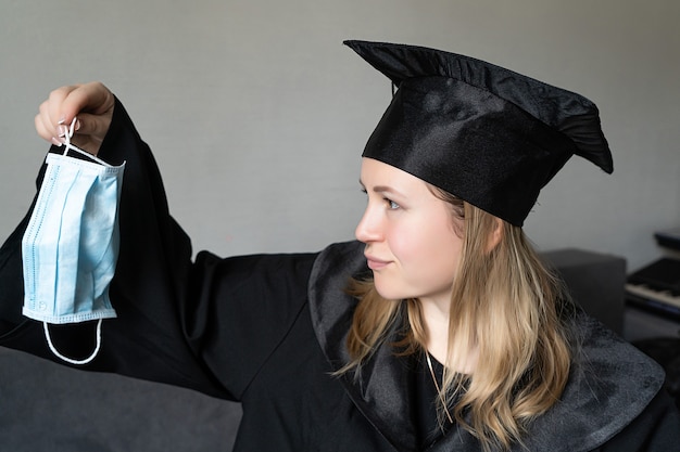 Fille tenant un masque médical avec un chapeau de graduation sur fond gris