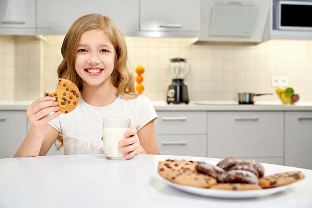 Fille tenant un verre de lait et des biscuits, posant dans la cuisine.