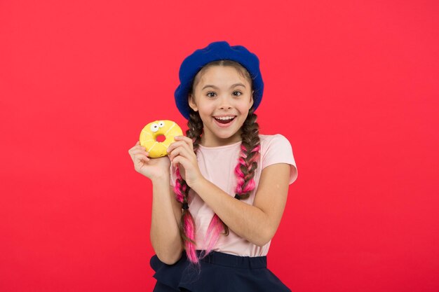 Fille tenir beignet mignon glacé à la main fond rouge Enfant fille souriante prête à manger un beignet Boutique de bonbons et concept de boulangerie Enfants grands fans de beignets cuits au four Impossible de résister à un beignet fraîchement préparé