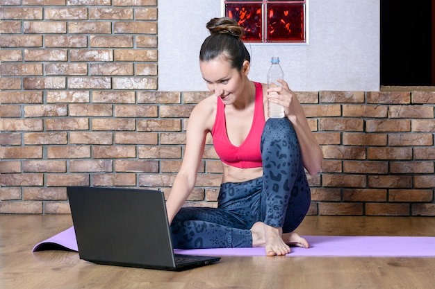 Une fille en tenue de sport fait une pause d'exercices en position assise sur le sol et regarde le réseau social de l'ordinateur portable avec une bouteille d'eau à la main