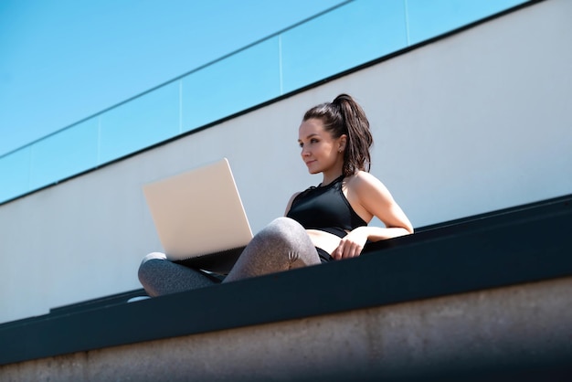 Une fille en tenue de sport sur un terrain de sport extérieur d'été est assise avec un ordinateur portable