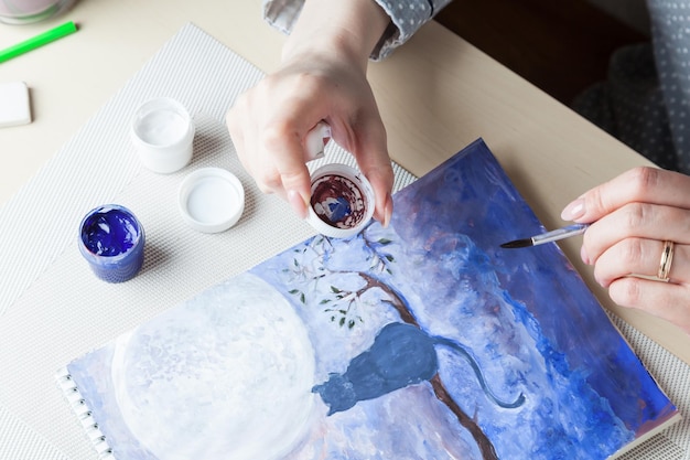 Photo une fille tient dans ses mains un pinceau et un couvercle d'un pot d'aquarelle l'arrière-plan d'un dessin d'un chat le ciel nocturne et la lune réalisés dans des tons bleus à proximité des outils occupation en mode quarantaine