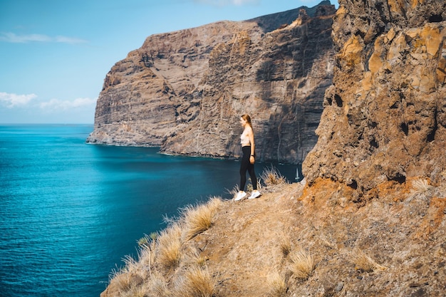 Une fille touristique calme et décontractée indépendante se dresse au bord d'une falaise avec une vue imprenable sur l'océan et le ciel Chaînes de montagnes de Los Gigantes Tenerife Santiago del Teide Îles Canaries Espagne