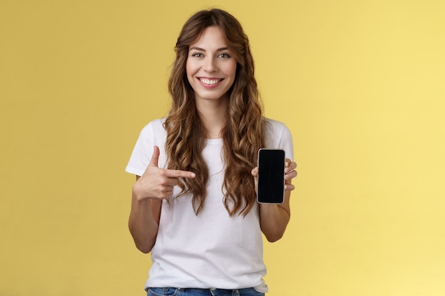 La fille a tout besoin d'un smartphone. Cheerful détendue femme insouciante longue coiffure bouclée tenir un téléphone portable pointant l'index téléphone portable souriant largement expliquer comment fonctionne l'application fond jaune