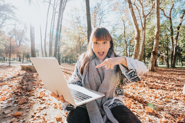 Fille travaillant sur ordinateur portable dans le parc en automne. Concept de travail indépendant. Heureuse jeune belle étudiante rouge ehad assise à l'extérieur dans un parc naturel à l'aide d'un ordinateur portable.Sourire à l'appareil photo heureux