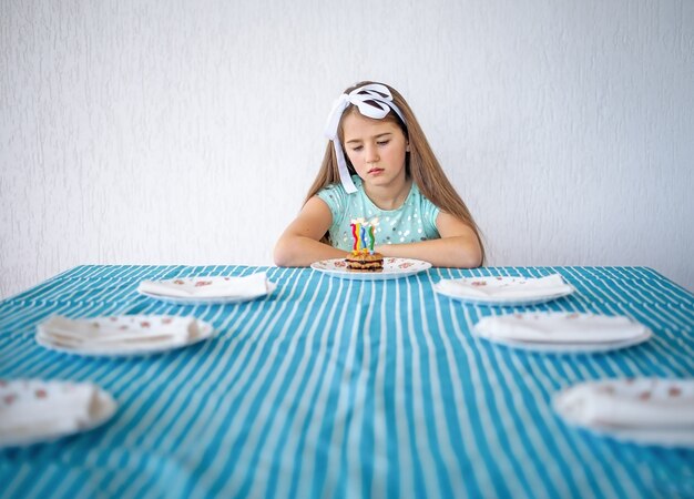 Une fille triste avec un gâteau avec des bougies est assise seule à une grande table. Concept de solitude.