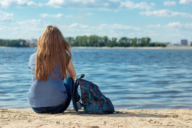 Une fille triste en jeans est assise sur le sable sur le talus avec un sac à dos dans le vent