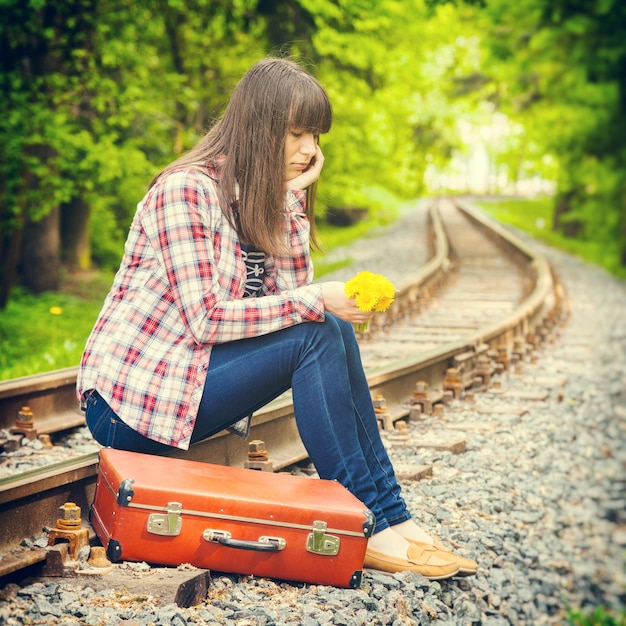 Fille triste avec valise et bouquet de pissenlits assis sur les rails