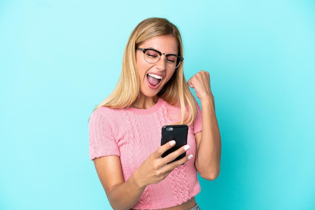 Fille uruguayenne blonde isolée sur fond bleu avec téléphone en position de victoire