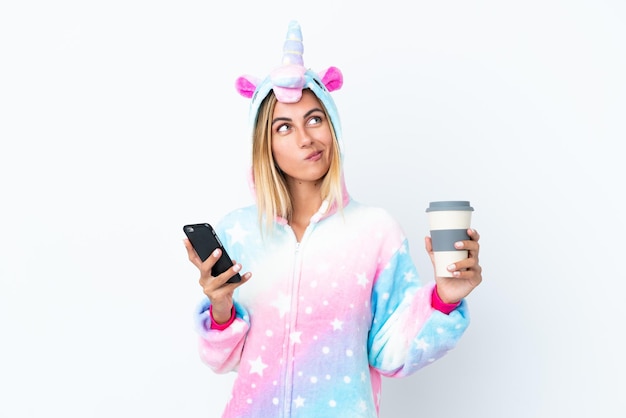 Fille uruguayenne blonde portant un pyjama licorne isolé sur fond blanc tenant du café à emporter et un mobile tout en pensant à quelque chose