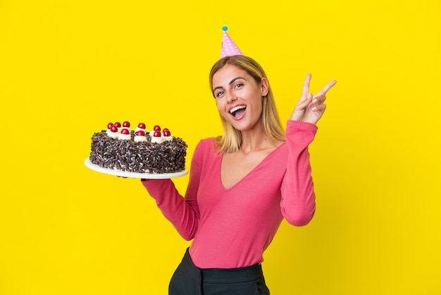 Fille uruguayenne blonde tenant un gâteau d'anniversaire isolé sur fond jaune souriant et montrant le signe de la victoire