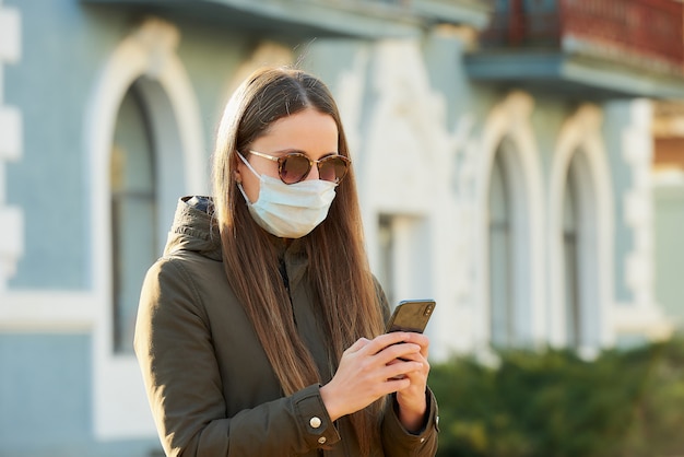 Une fille utilisant un smartphone porte un masque médical pour éviter la propagation du coronavirus dans une rue de la ville. Une femme à lunettes de soleil avec un masque chirurgical sur le visage contre COVID-19.