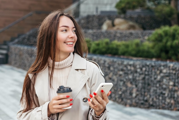 fille utilise son smartphone tient une tasse de café et sourit dans le contexte d'un immeuble de bureaux