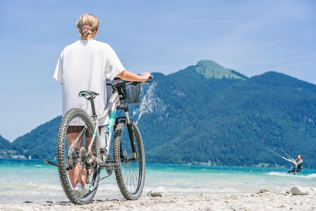 Une fille avec un vélo de montagne se tient sur la rive du lac et regarde les montagnes.