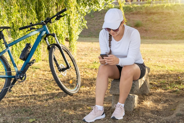 Une fille vérifie son téléphone portable et se repose sous un arbre à côté de son vélo après avoir fait de l'exercice