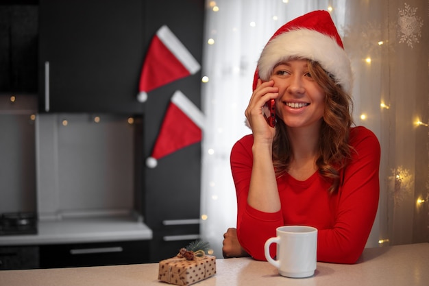 Fille en vêtements rouges dans la cuisine parlant sur un smartphone la veille de Noël