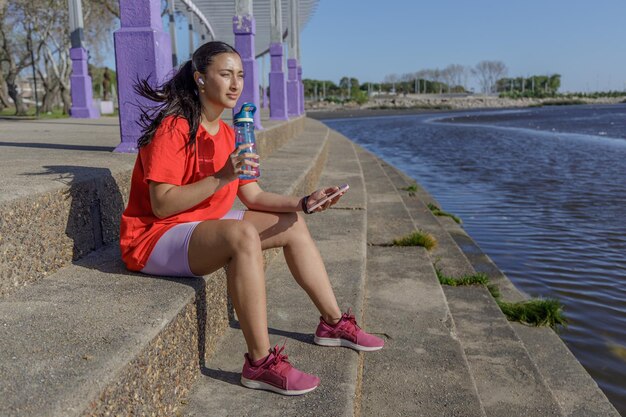 Une fille en vêtements de sport assise sur les escaliers buvant dans une bouteille d'eau.