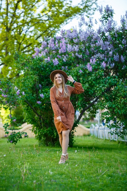 Une fille vêtue d'un chapeau et d'une robe marron se promène dans le parc, elle a une apparence européenne et un sourire sur son visage. La jeune femme attirante marche un jour ensoleillé