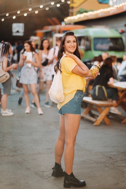 Une fille vêtue d'une chemise jaune, d'un short et de chaussures noires avec un filet de citrons et de jus de citron se promène dans une rue de la ville. Une femme avec un cocktail dans ses mains.