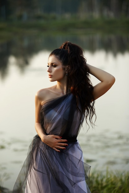 Fille vêtue d'une longue robe près du lac est assise sur un vieux bateau, une belle femme dans les rayons du soleil du soir à l'automne dans le village. Vie rurale et mode