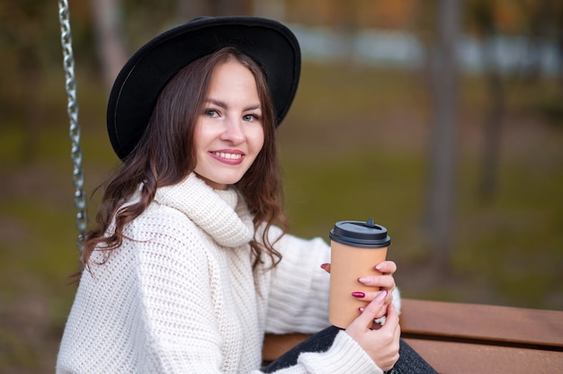 Une fille vêtue d'un pull blanc et d'un chapeau noir à l'automne est assise sur un banc. Tient un verre de café dans ses mains.