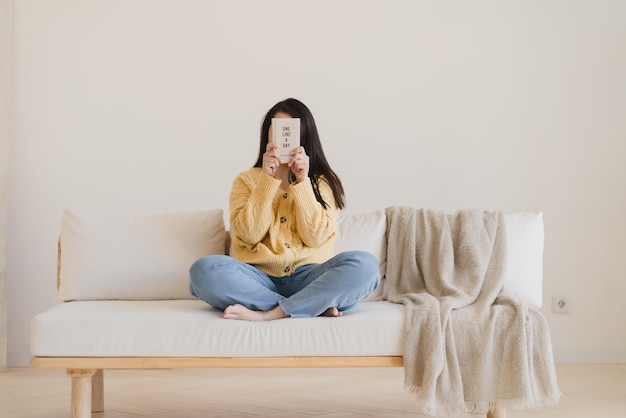 Une fille vêtue d'un pull jaune et d'un jean bleu est assise sur un canapé et se couvre le visage avec un livre