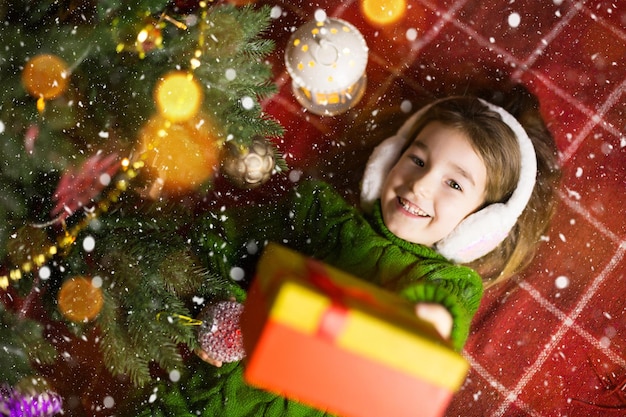 Une fille vêtue d'un pull en tricot chaud et d'un casque en fourrure est allongée sur une couverture près de l'arbre de Noël avec une boîte-cadeau. Le réveillon de Noël, un rêve et un souhait. Nouvel An, ambiance festive, guirlandes et bokeh