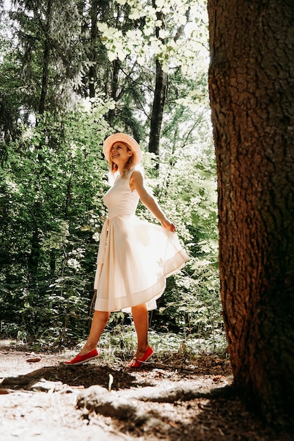 Une fille vêtue d'une robe blanche et d'un chapeau se promène dans un parc ou une forêt d'été. Forêt de fées. Photo pleine longueur d'une femme heureuse