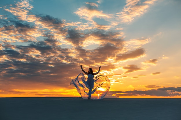 Une fille vêtue d'une robe blanche volante danse et pose dans le désert de sable au coucher du soleil