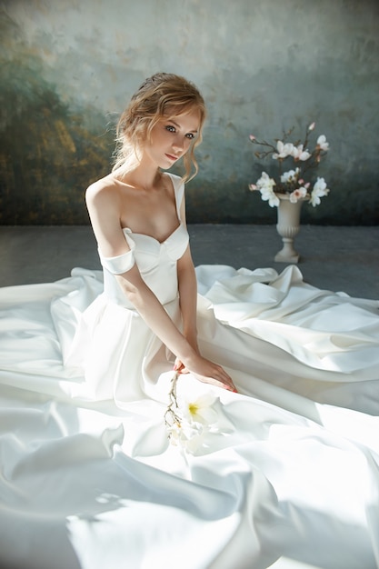 Fille vêtue d'une robe longue chic assise sur le sol. Robe de mariée blanche sur le corps de la mariée. Belle robe légère avec un long ourlet, portrait d'une jeune fille fragile et délicate avant la cérémonie de mariage