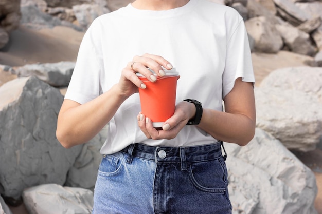 Photo une fille vêtue d'un t-shirt blanc et d'un jean bleu tient une tasse rouge pour un café à emporter dans le contexte d'une plage de mer avec du sable et des rochers gris