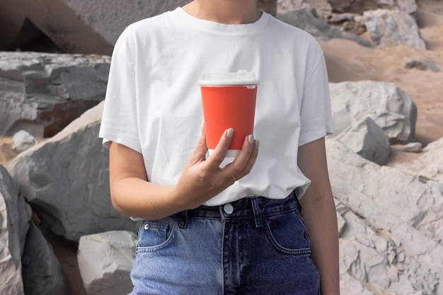 Photo une fille vêtue d'un t-shirt blanc et d'un jean bleu tient une tasse rouge pour un café à emporter dans le contexte d'une plage de mer avec du sable et des rochers gris