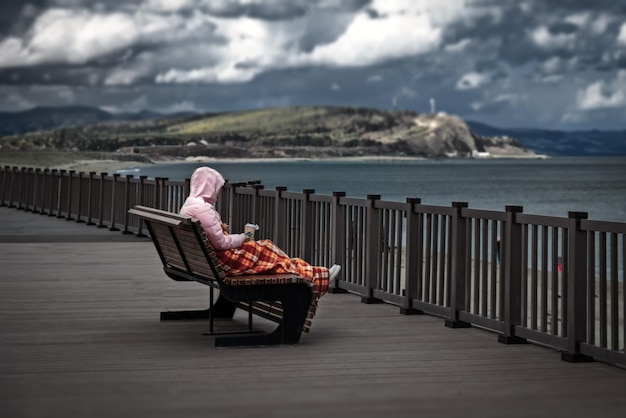 Une fille vêtue d'une veste enveloppée d'une couverture est assise avec une tasse de café sur le banc sur la rive de la mer froide et sombre.