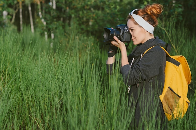 Une fille vêtue d'une veste grise avec un sac à dos jaune tient une caméra photo-vidéo professionnelle dans ses mains. Dans le contexte de la nature verte et de la forêt.