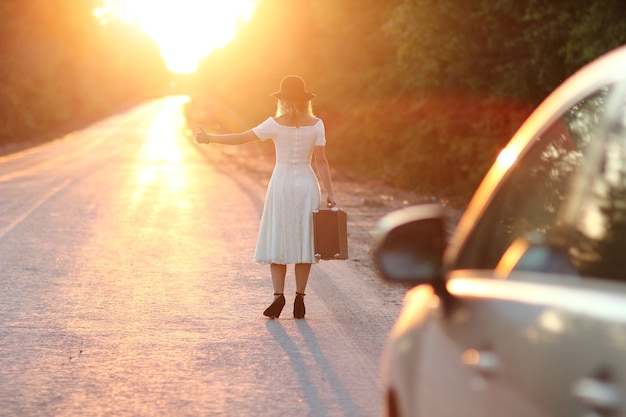 Fille vintage avec chapeau avec une valise faisant de l'auto-stop en voiture