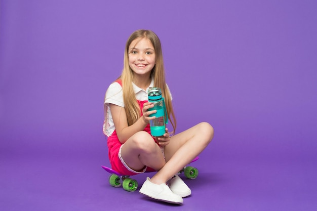 Une fille sur un visage souriant tient avec une bouteille d'eau tout en étant assise sur un fond violet de planche à roulettes. Une fille aux cheveux longs boit de l'eau.