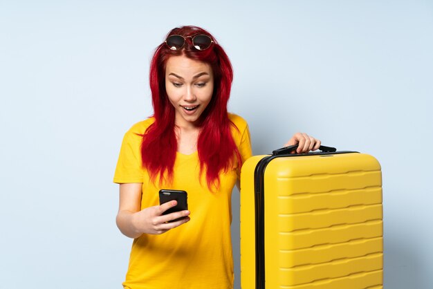 Fille de voyageur tenant une valise isolée sur bleu surpris et en envoyant un message