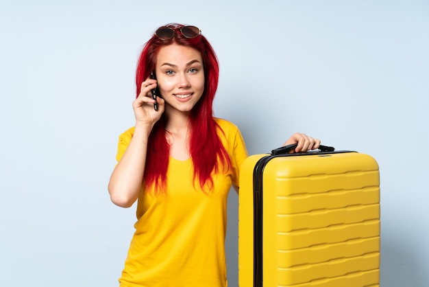 Fille de voyageur tenant une valise isolée sur le mur bleu en gardant une conversation avec le téléphone mobile avec quelqu'un