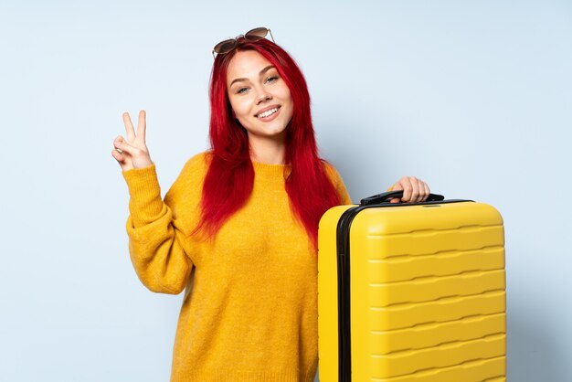 Fille de voyageur tenant une valise sur le mur bleu souriant et montrant le signe de la victoire