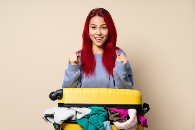 Fille de voyageur avec une valise pleine de vêtements isolé sur fond beige célébrant une victoire en position de gagnant