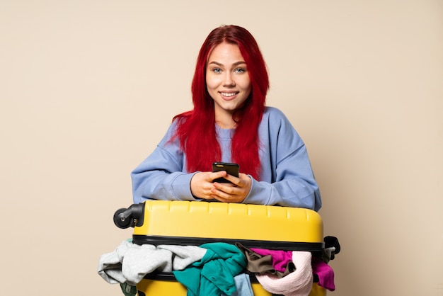 Fille de voyageur avec une valise pleine de vêtements isolés sur beige en envoyant un message avec le mobile