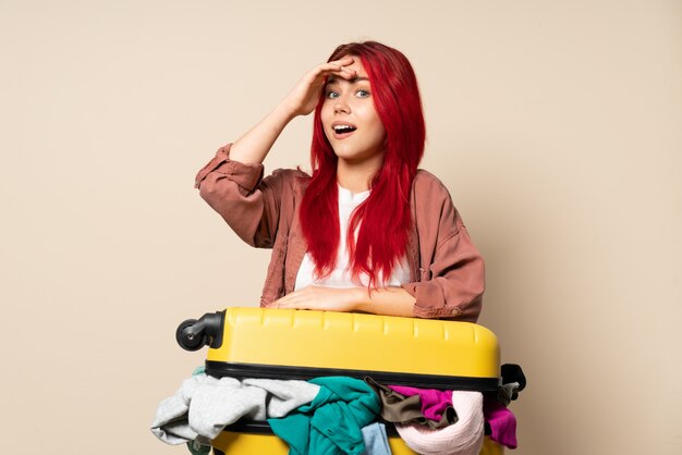 Fille de voyageur avec une valise pleine de vêtements isolés sur beige faisant un geste surprise tout en regardant sur le côté