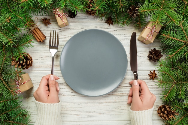 La fille vue de dessus tient une fourchette et un couteau à la main et est prête à manger. Assiette vide ronde en céramique sur Noël en bois. plat de dîner de fête avec décor de nouvel an