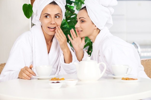 Filles au spa. Deux belles jeunes femmes en peignoir buvant du thé et bavardant assis devant la piscine